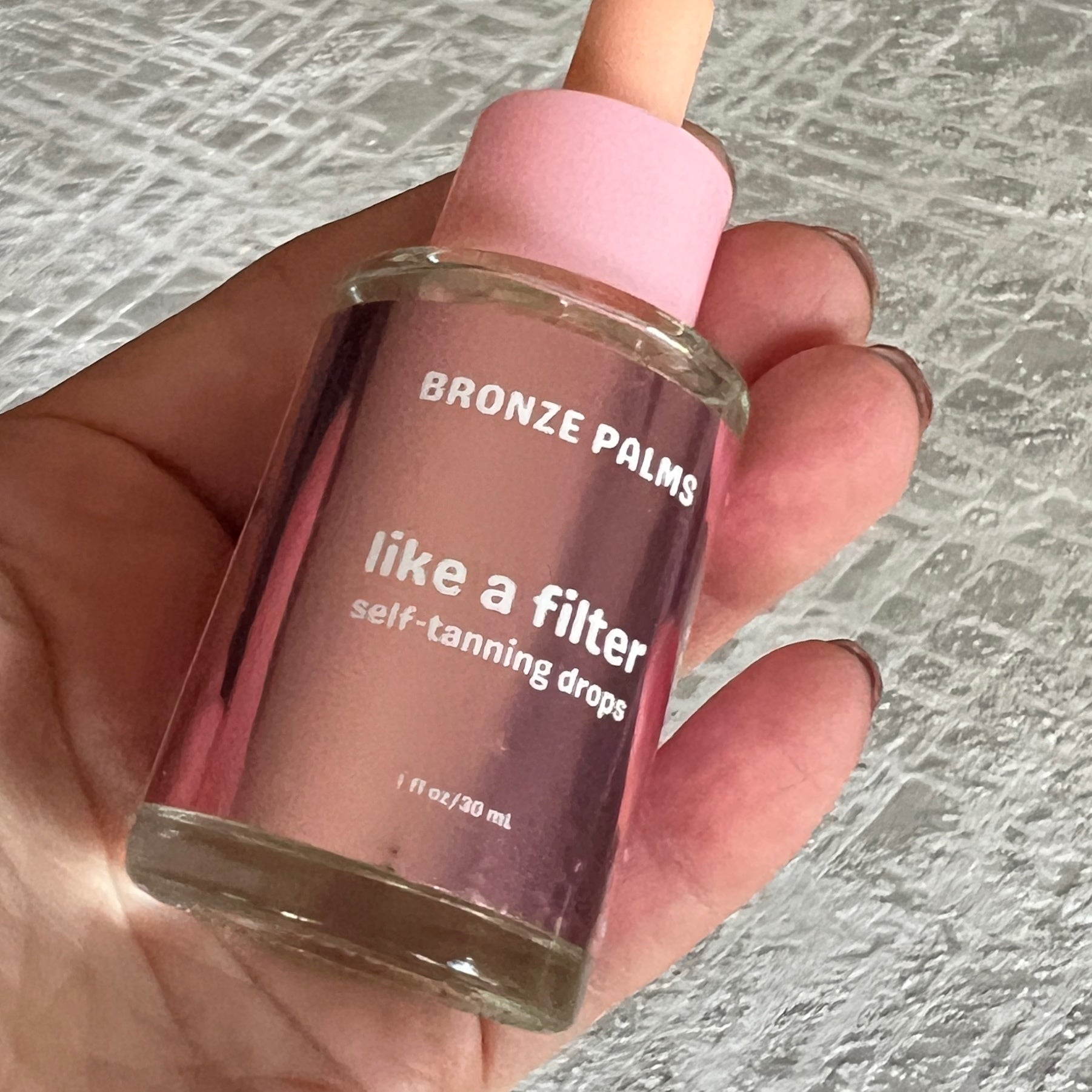 Hand holding a bottle of 'Bronze Palms like a filter self-tanning drops' with a pink cap.